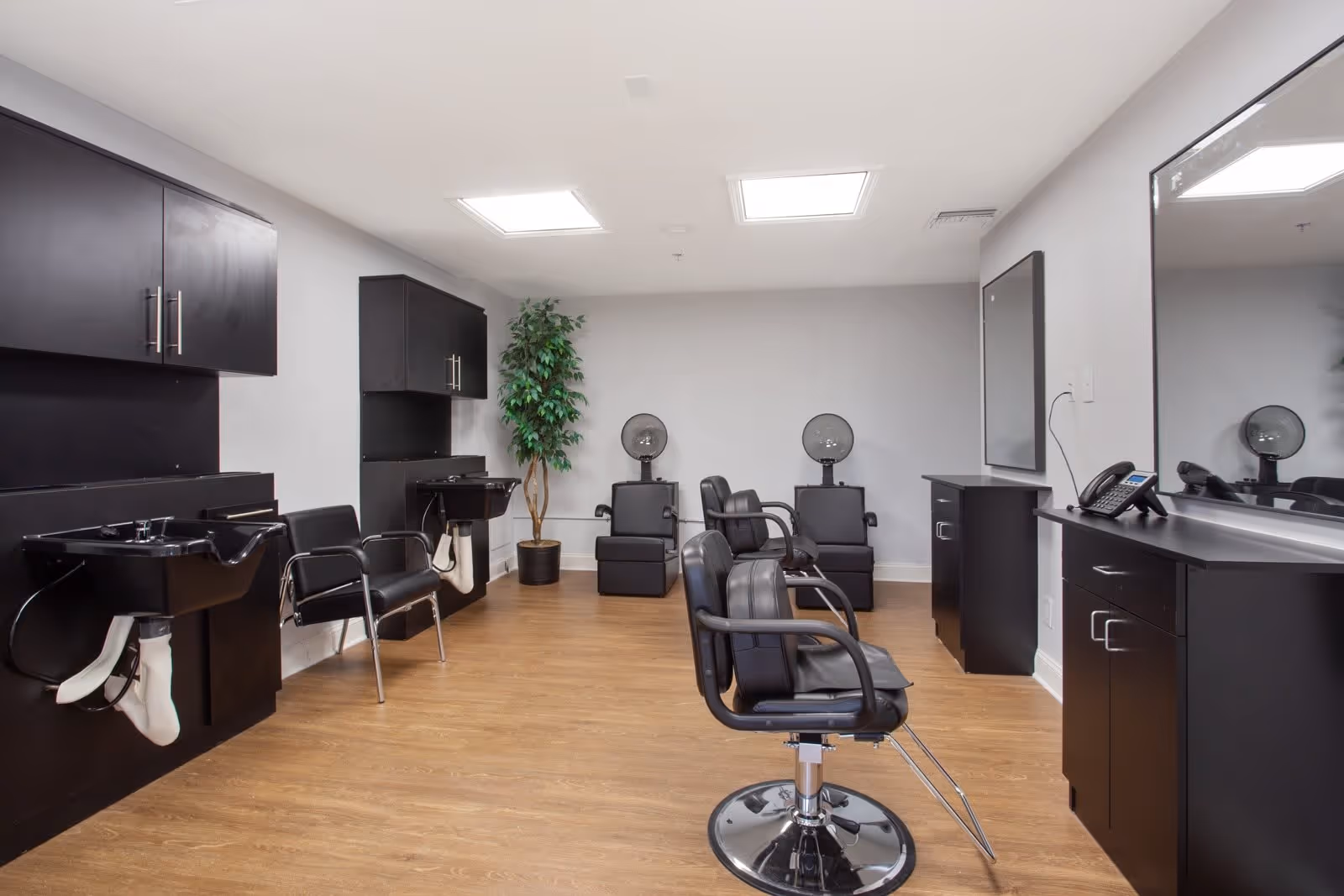 Interior view of a hair salon area in a senior living facility with black salon chairs, hair washing stations, hair dryers, black cabinetry, a large mirror, and a potted plant against light gray walls and wood flooring.
