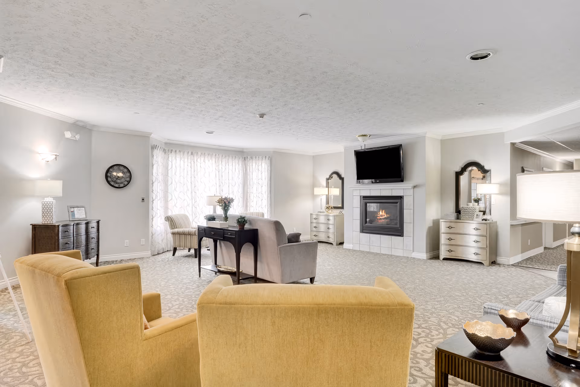 Spacious bright living room lounge with upholstered chairs, a fireplace with a TV above, side tables and lamps.