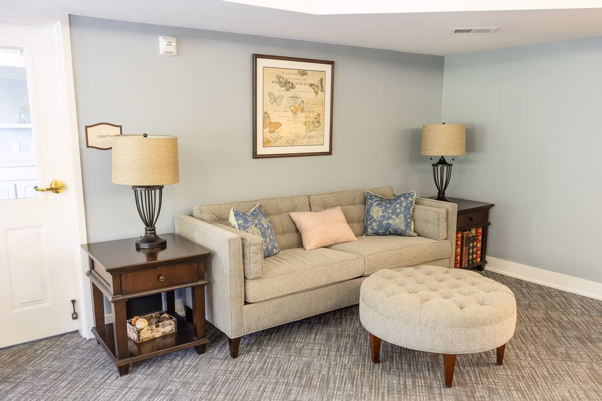Neutral sitting area with a gray sofa, patterned throw pillows, a round tufted ottoman, two side tables with lamps, and framed art on a pale blue wall.