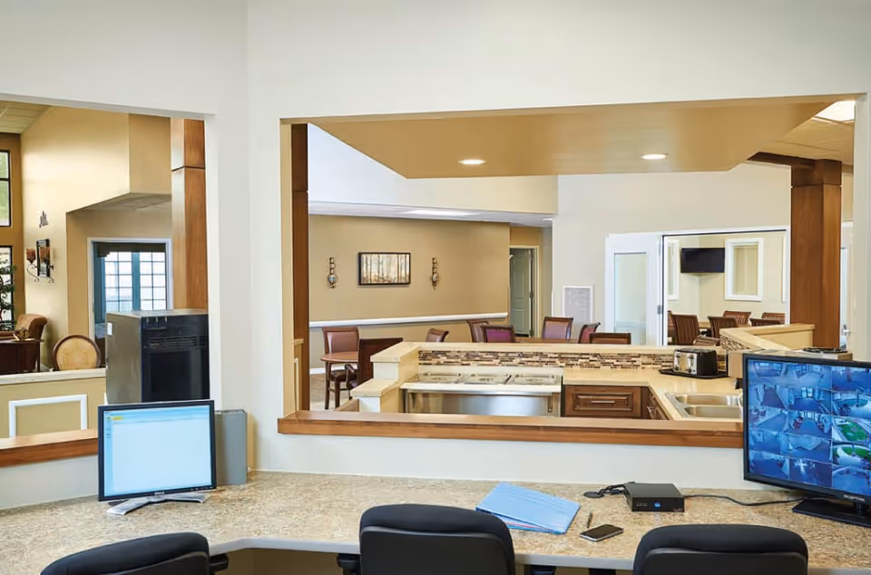 Interior view of a senior living facility common area showing a reception desk with computer monitors facing an open kitchen and dining seating.