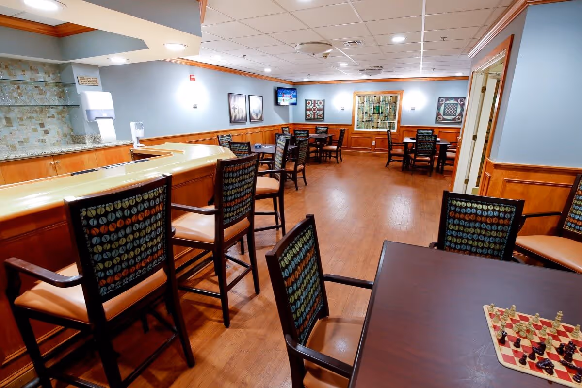A spacious common area with wooden flooring and light blue walls featuring wood paneling. There are several tables and chairs arranged throughout the room, including a table with a chessboard and chess pieces set up. A counter with high chairs is on the left side, and a small wall-mounted TV is visible in the background. The room is well-lit with ceiling lights and decorated with framed artwork on the walls.