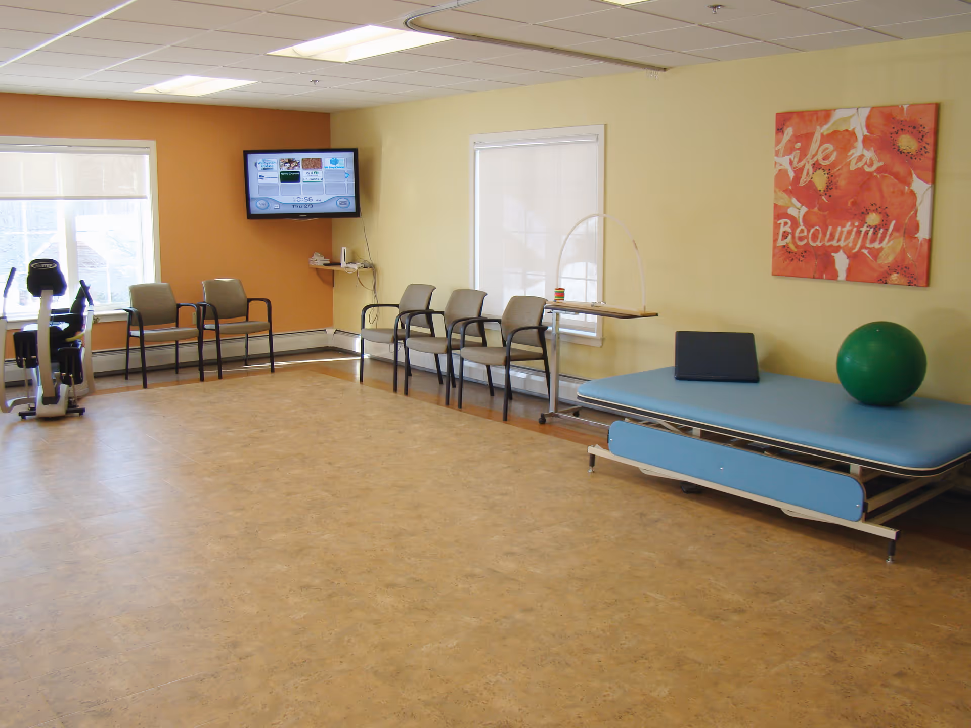 A rehabilitation room with exercise equipment including a stationary bike, several chairs lined up against the walls, a blue therapy table with a green exercise ball and a black cushion on it, a wall-mounted TV, and a colorful wall art that says 'Life is Beautiful'.