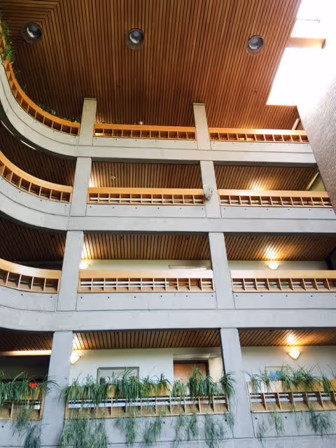 Interior view of a multi-story building atrium with concrete balconies and wooden ceiling panels. Each floor has railings with plants placed along the edges, and there are wall-mounted lights illuminating the space.