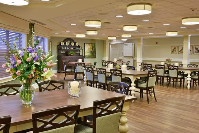 A spacious dining room with multiple wooden tables and chairs arranged neatly. A large vase with a colorful bouquet of flowers sits on the nearest table, along with a glass candle holder. The room has wooden flooring, beige walls, and ceiling lights. Windows with blinds are on the left side, and a dark wooden cabinet with decorative plates and a framed painting are visible in the background.