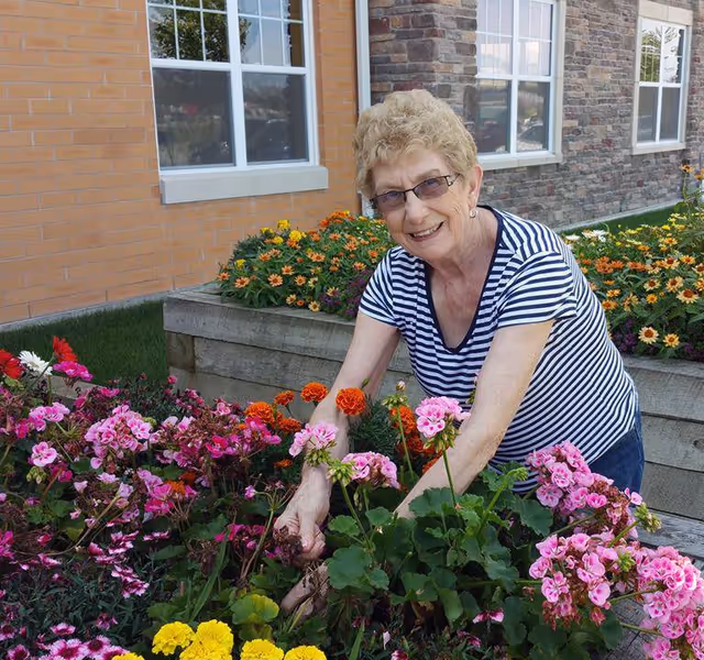 An elderly woman wearing glasses and a striped shirt is smiling while tending to a colorful flower garden outside a building with brick and stone walls and white-framed windows.