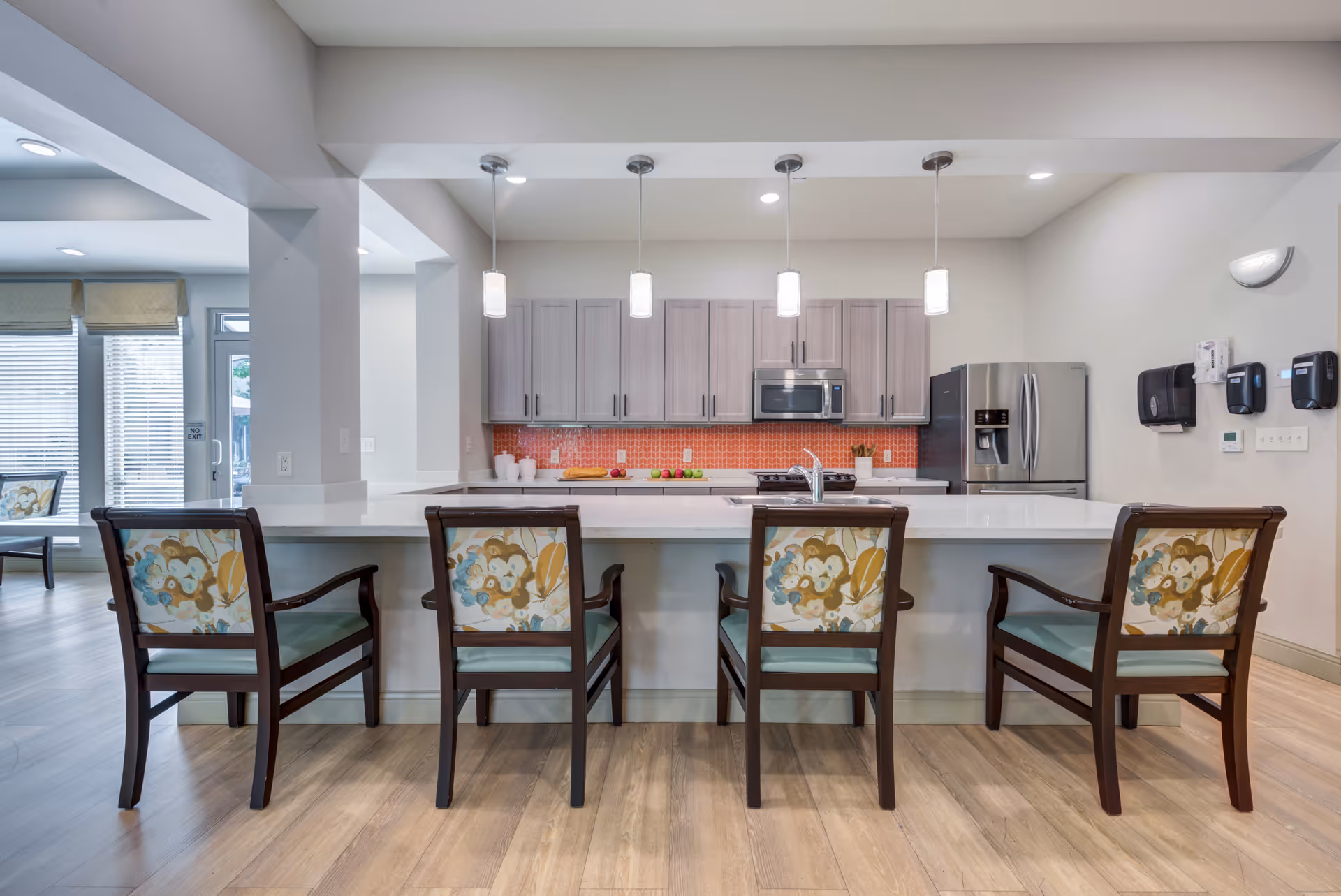 Communal kitchen with a long white island lined with four patterned chairs, pendant lights, stainless steel appliances and gray cabinets.