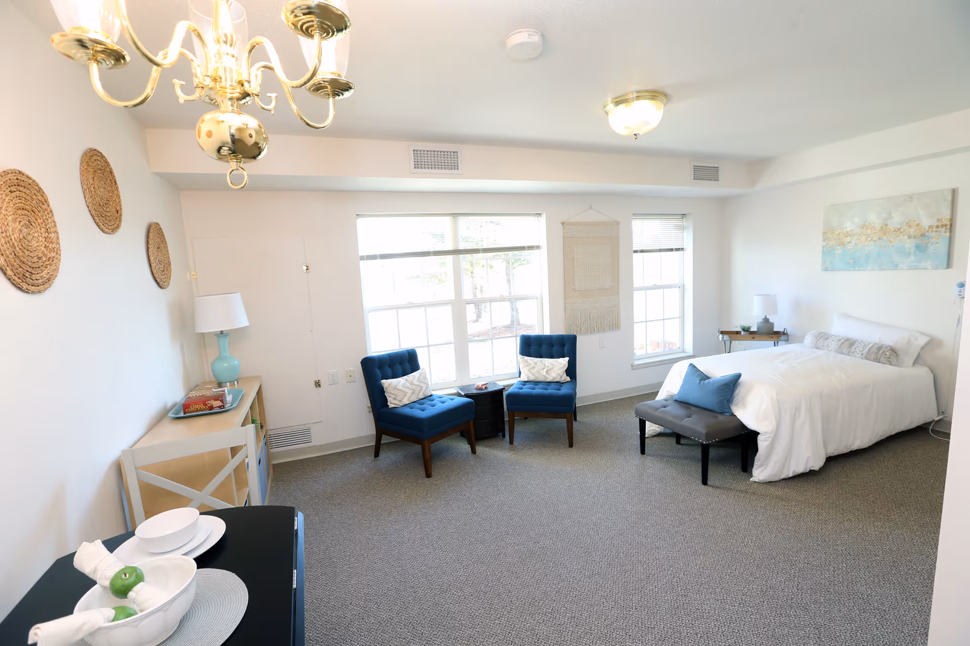 A bright and spacious bedroom in a senior living facility with a white bed, blue accent pillows, two blue upholstered chairs with white cushions, a small black side table, a bench at the foot of the bed, a console table with a lamp and books, and decorative wall hangings including woven baskets and a painting. The room has large windows allowing natural light to fill the space.