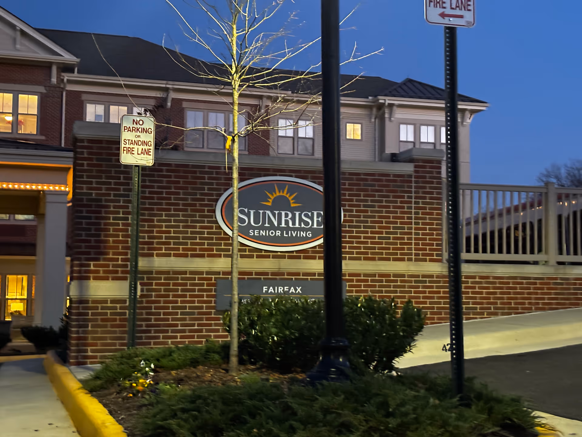 Exterior view of Sunrise Senior Living facility in Fairfax at dusk, showing a brick wall with the Sunrise Senior Living sign and a small tree in front. There are two signs indicating no parking or standing in the fire lane.