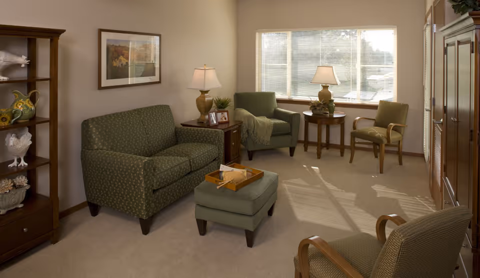 A cozy living room with a green patterned loveseat, matching ottoman, and three armchairs. There is a wooden side table with a lamp and framed photos, a round table with a lamp and plant near a large window with blinds, and a wooden shelving unit with decorative items on the left side.