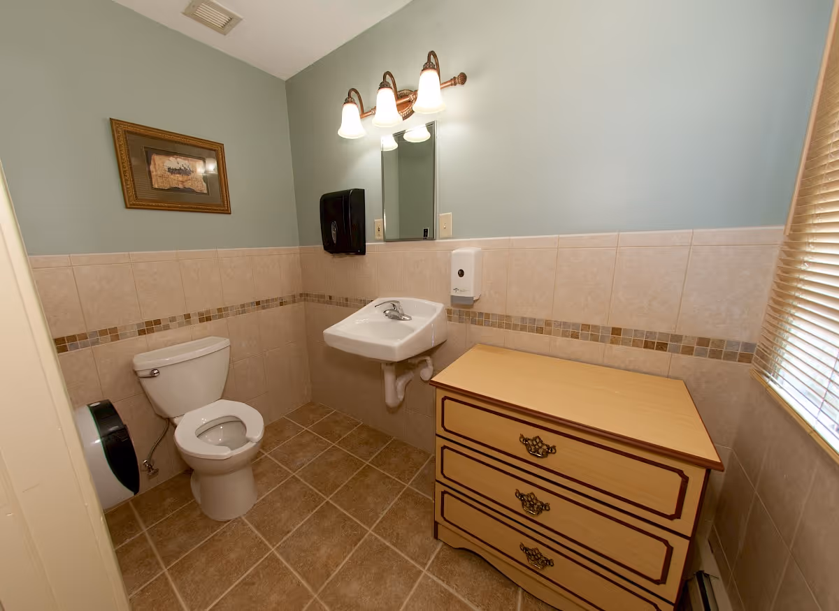 A clean bathroom with beige tiled walls and floor, featuring a white toilet, a wall-mounted white sink with a mirror and three-light fixture above it, a wooden dresser with three drawers, a wall-mounted soap dispenser, a paper towel dispenser, a framed picture on the wall, and a window with closed blinds.