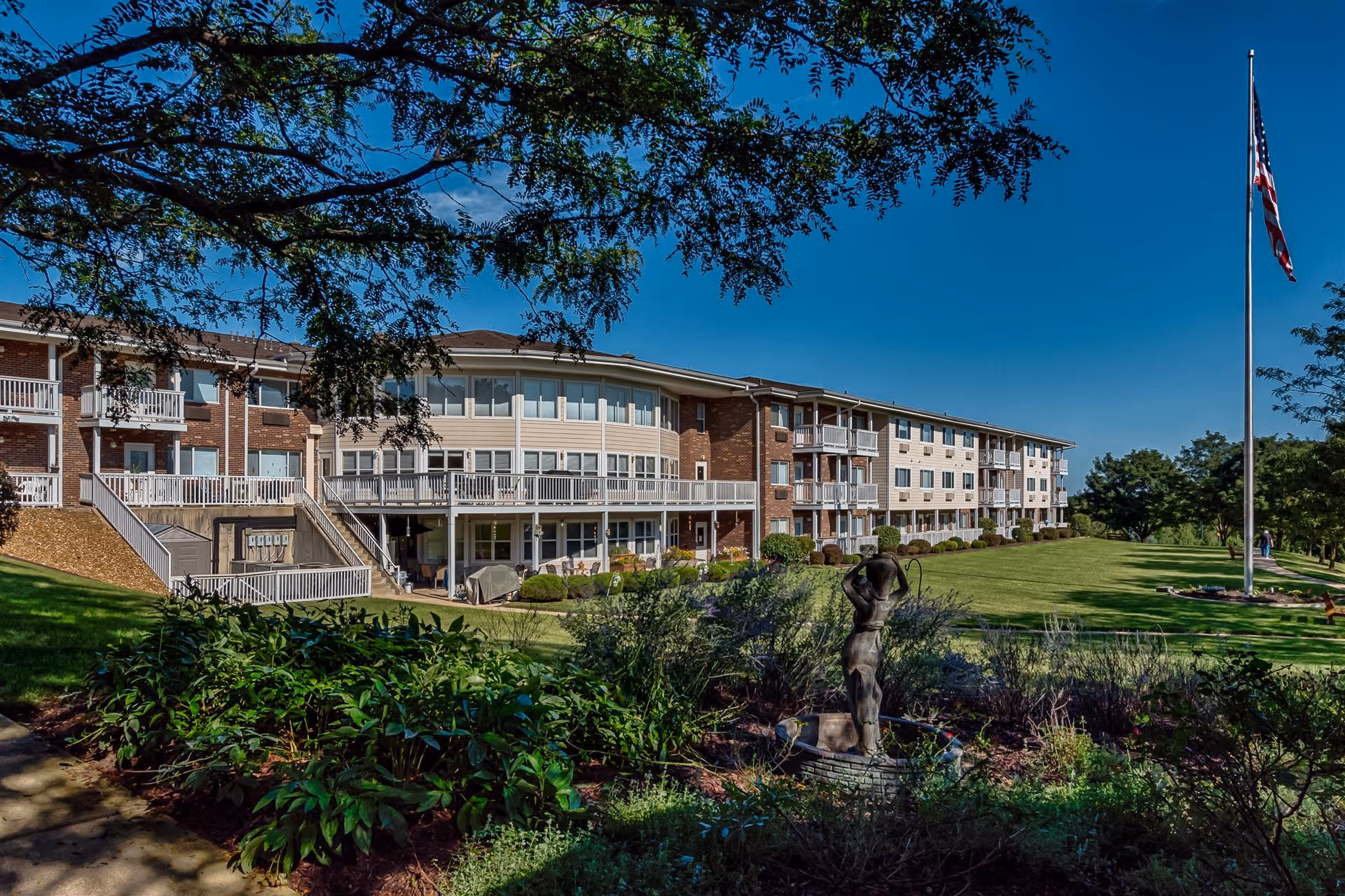 Front view of a multi-story assisted living building with balconies, landscaped lawn, flagpole, and a small statue in the foreground.