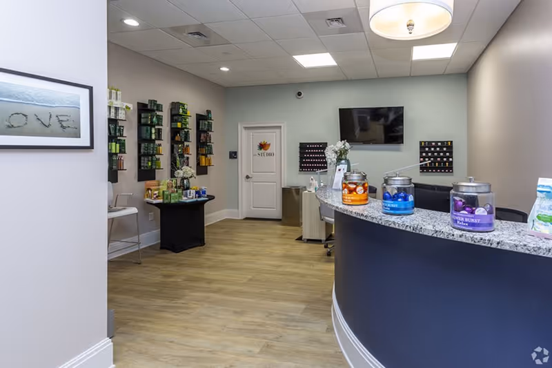 Reception area with a curved granite countertop displaying jars of colorful bath salts, a wall-mounted TV, a door labeled 'Studio', shelves with various products, and a framed picture on the wall that says 'LOVE'.