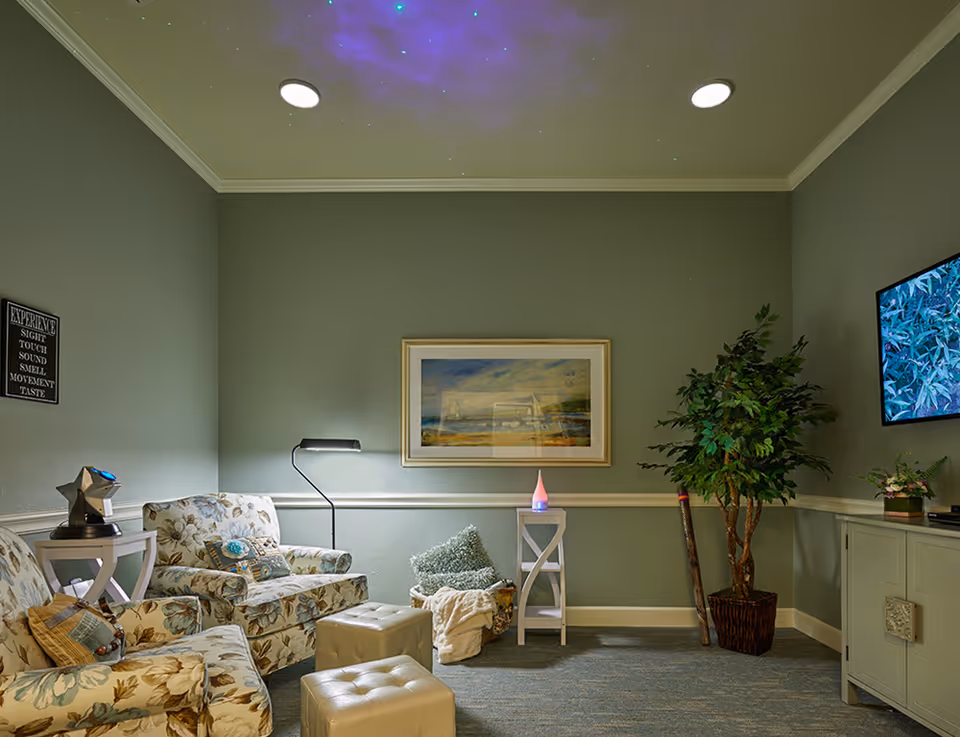 A cozy living room with floral patterned armchairs, a small white side table with a star-shaped device, a floor lamp, a framed landscape painting on the wall, a potted plant, a TV mounted on the wall displaying foliage, and a small white cabinet. The ceiling has a purple light projection resembling a night sky.