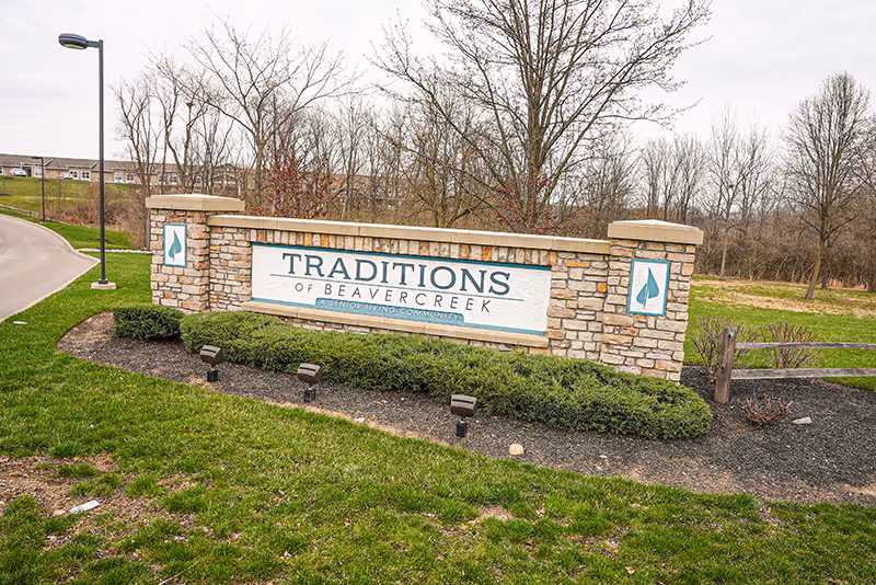 Stone entrance sign reading "TRADITIONS OF BEAVERCREEK" set in landscaped grass with trees and a nearby streetlamp.