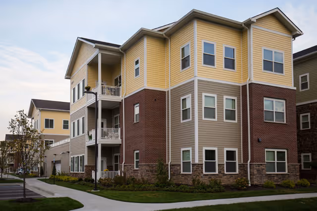 Exterior view of a multi-story residential building with yellow, beige, and brick siding, surrounded by landscaped greenery and a sidewalk.
