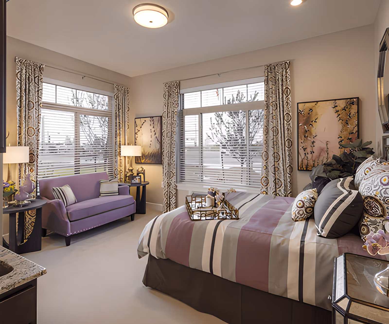 A well-lit bedroom with a large bed featuring striped bedding in shades of purple, gray, and white. The room has two large windows with patterned curtains, a purple loveseat with two pillows, two side tables with lamps, and decorative artwork on the walls. A decorative tray with flowers and items is placed on the bed.