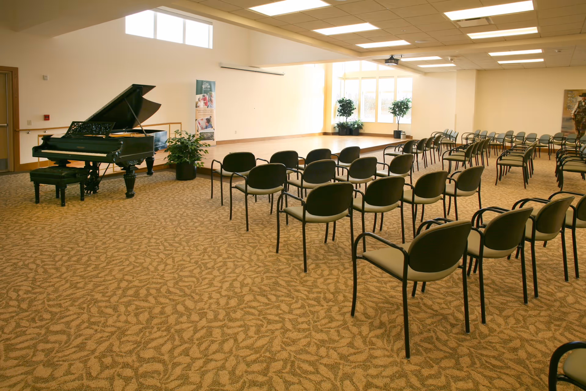 A spacious room with a grand piano on the left side and rows of chairs arranged facing a small stage area with large windows and potted plants in the background. The room has patterned carpet and ceiling lights.