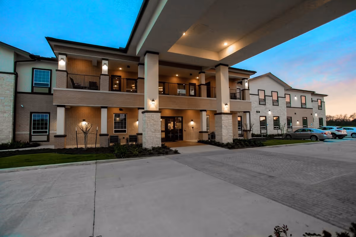 Exterior view of a two-story senior living facility building at dusk with lights on, a covered entrance supported by columns, balconies, and a parking area with several cars.