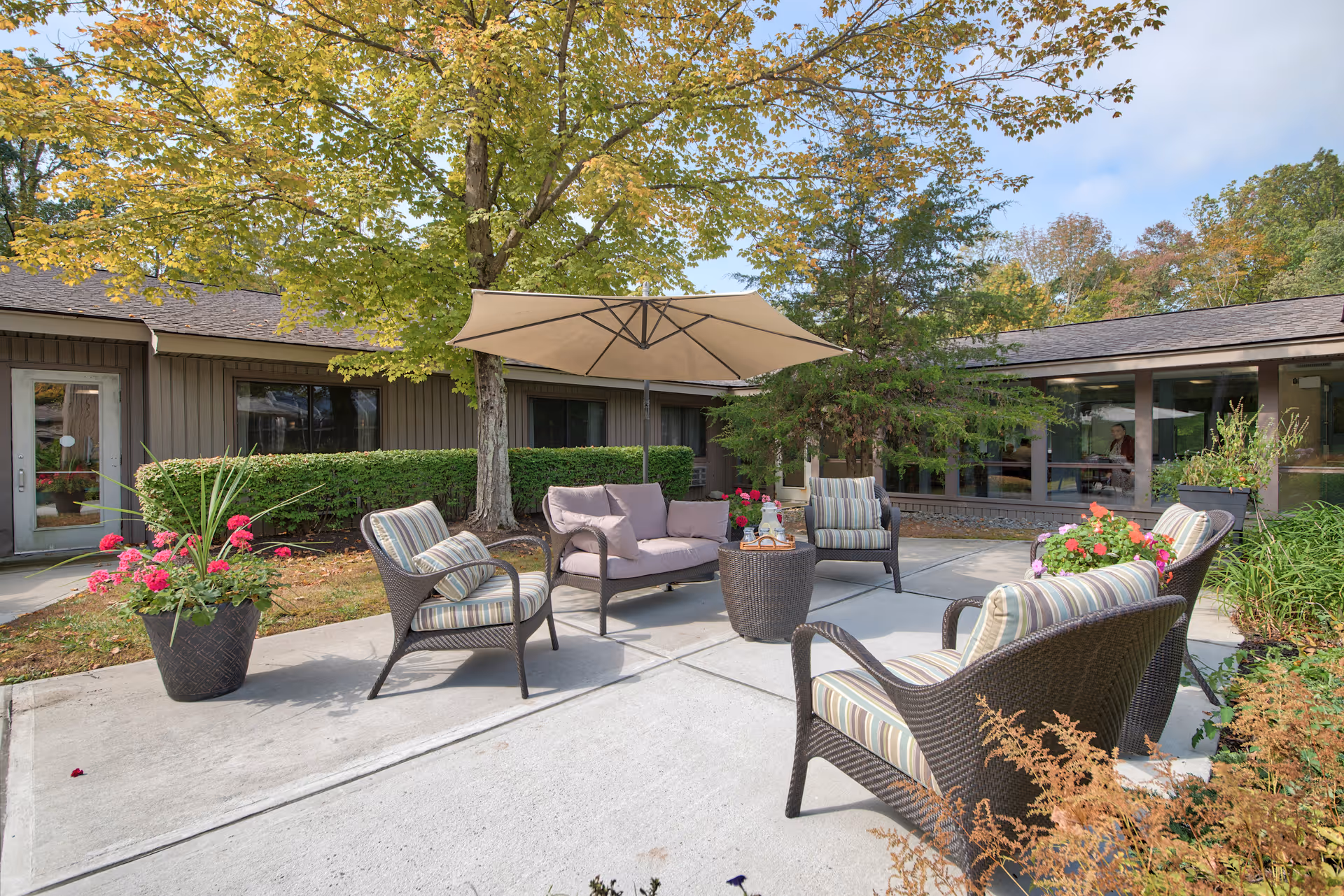 Outdoor patio area at The Country House in Westchester with cushioned wicker chairs and a loveseat arranged around a small round table. A large beige umbrella provides shade, and there are potted plants with flowers around the seating area. The patio is surrounded by trees and shrubs, with a building visible in the background.