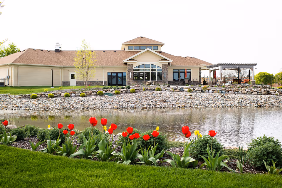 Exterior view of a single-story senior living facility building with beige siding and a brown roof, situated behind a small pond with a rocky shoreline. In the foreground, there is a well-maintained garden with red and yellow tulips and green grass. To the right of the building, there is a pergola with outdoor seating.