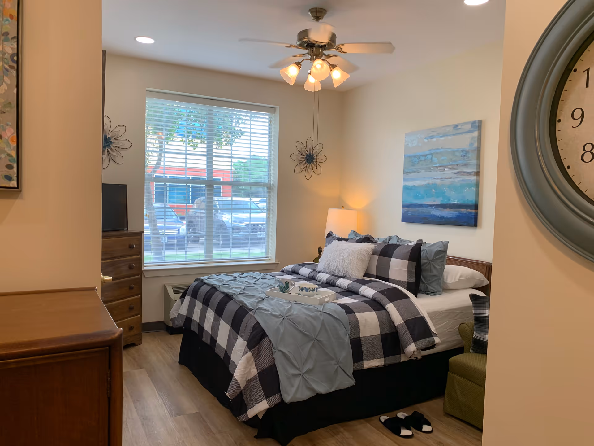 A cozy bedroom with a large window letting in natural light. The bed is made with a black and white checkered comforter, blue pillows, and a fluffy white accent pillow. A ceiling fan with lights hangs above the bed. There is a wooden dresser with a TV on top near the window, a green armchair beside the bed, and a large clock on the wall. A painting with blue and white tones is hung above the bed, and slippers are placed on the wooden floor near the bed.