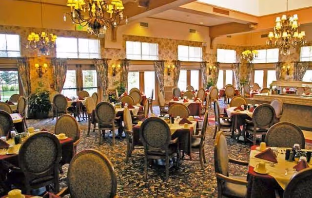 Spacious formal dining room with multiple round tables, upholstered chairs, chandeliers and large windows with curtains.
