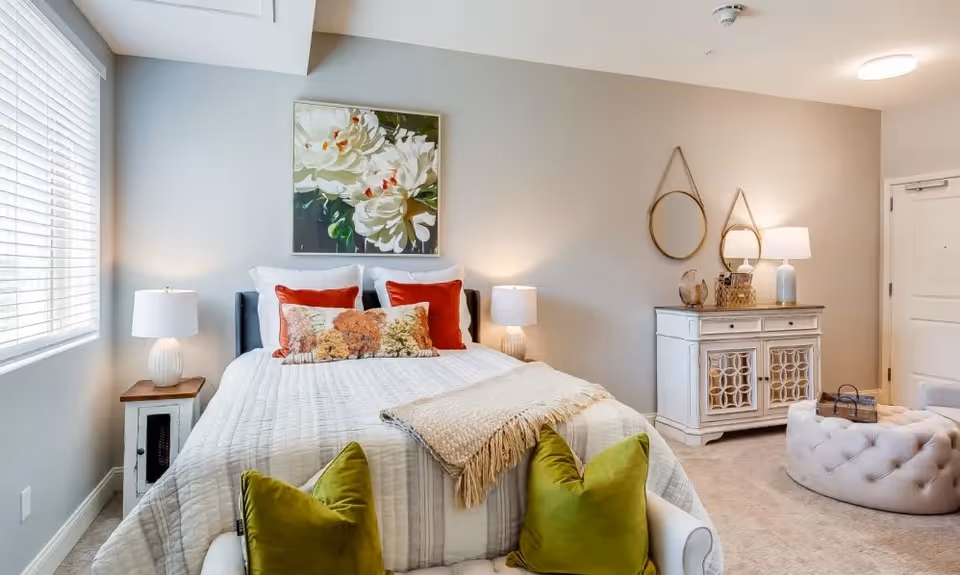 A cozy bedroom with a neatly made bed featuring white bedding, two red pillows, and a decorative pillow with a nature scene. Two green pillows are placed on a bench at the foot of the bed. The room has a large window with blinds on the left, two bedside tables with lamps, a white cabinet with decorative items and a lamp on the right, and two round mirrors hanging on the wall above the cabinet. The walls are painted light beige and the floor is carpeted.