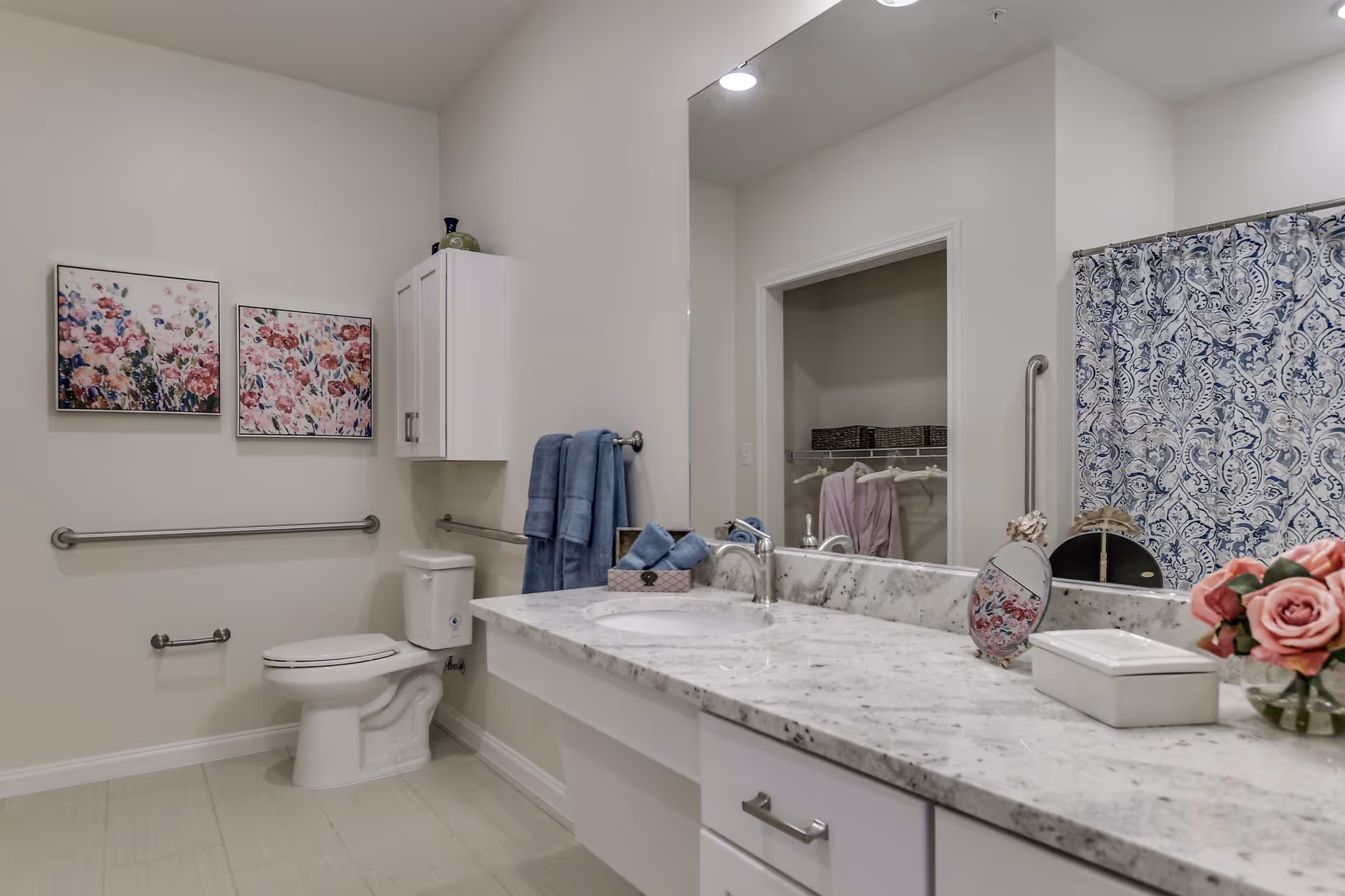 A clean and modern bathroom with a white toilet, a marble countertop with a sink, blue towels hanging on a rack, floral artwork on the wall, and a shower with a blue and white patterned curtain. There is a large mirror above the sink reflecting part of a closet with hanging robes and baskets.