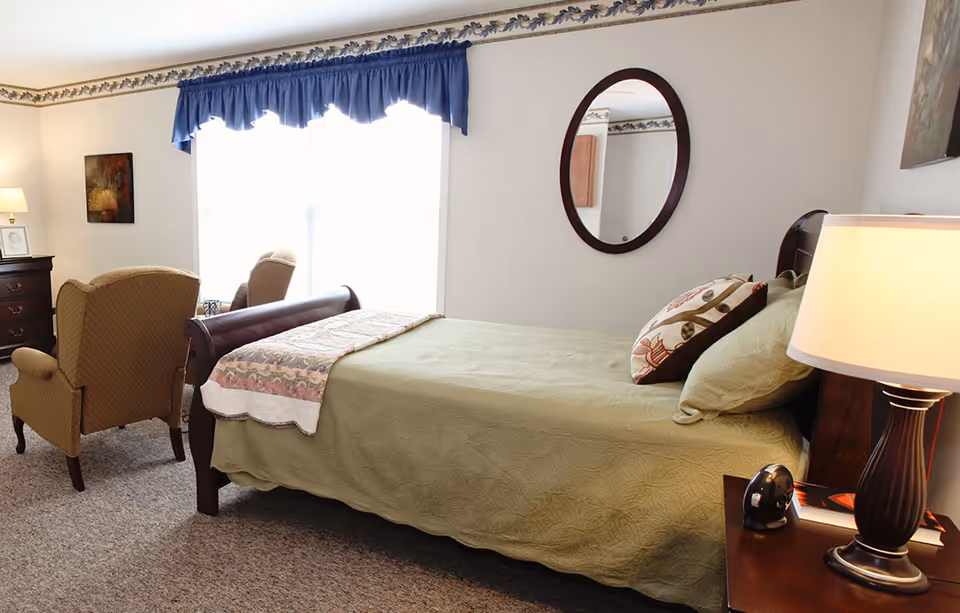 Light-filled bedroom with a single bed, nightstand lamp, two upholstered chairs and a dresser under a window with a blue valance.
