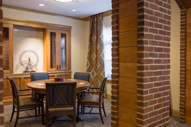 Round wooden dining table with six chairs in a cozy room framed by brick pillars and built-in cabinetry.