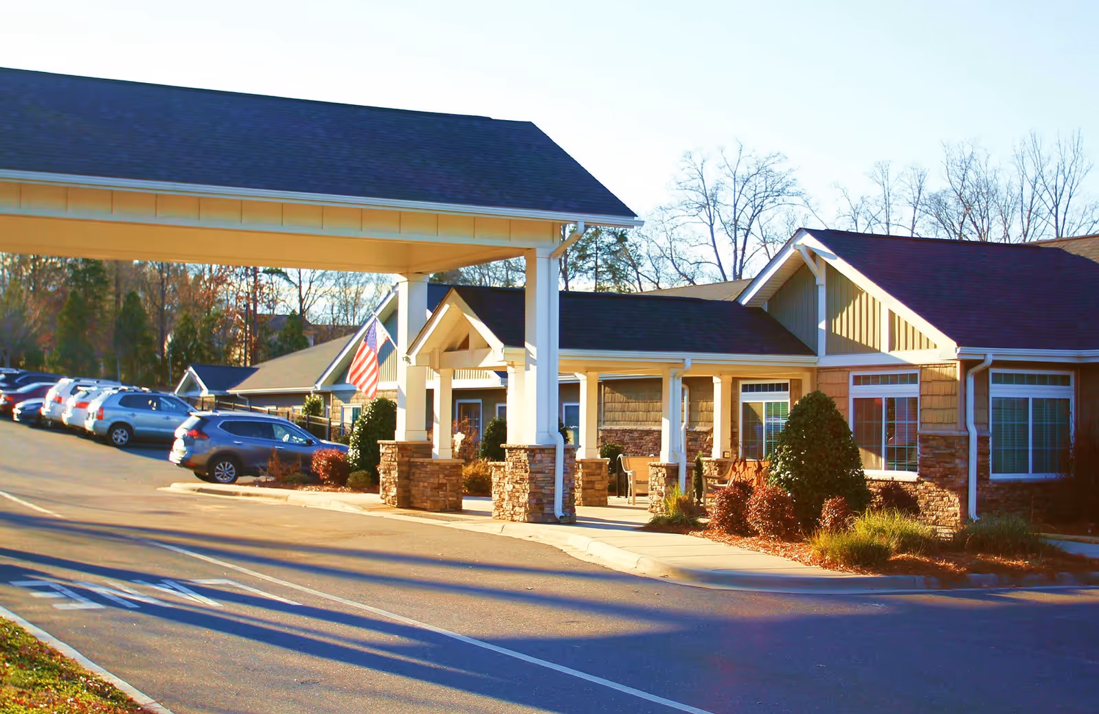 Exterior view of a senior living facility entrance with a covered drop-off area supported by white columns with stone bases. There are several parked cars along the driveway, and the building features beige siding, large windows, and landscaped bushes. An American flag is displayed near the entrance.