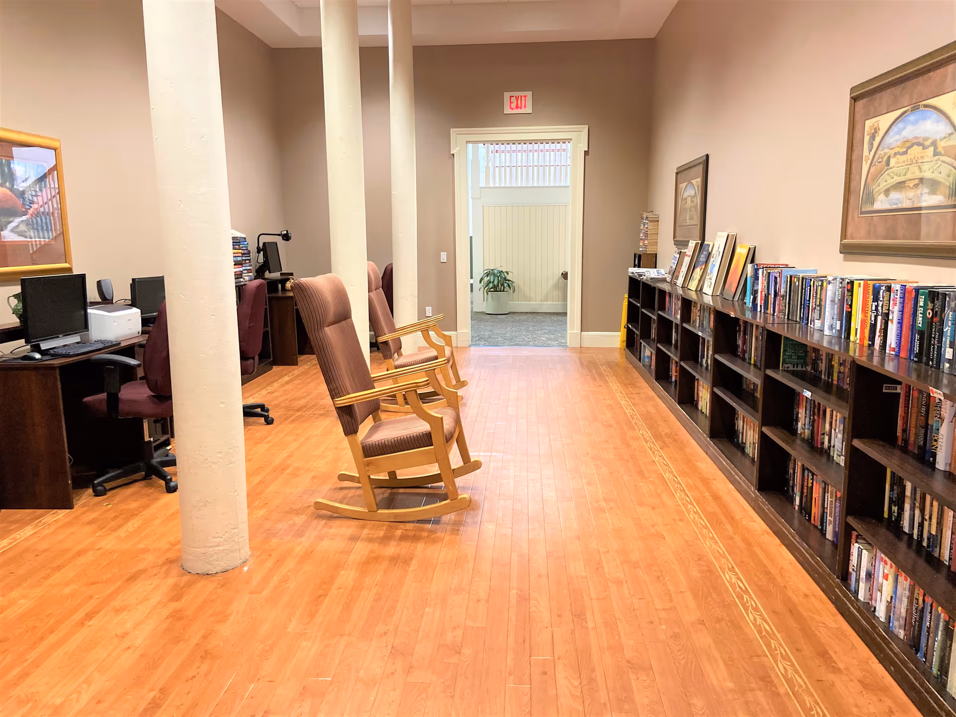 Interior view of a quiet room in Whaler's Cove Assisted Living featuring wooden flooring, two wooden rocking chairs with cushions, a long bookshelf filled with books along the right wall, framed artwork above the bookshelf, and computer desks with chairs along the left wall. A doorway with an exit sign above it is visible at the far end of the room.