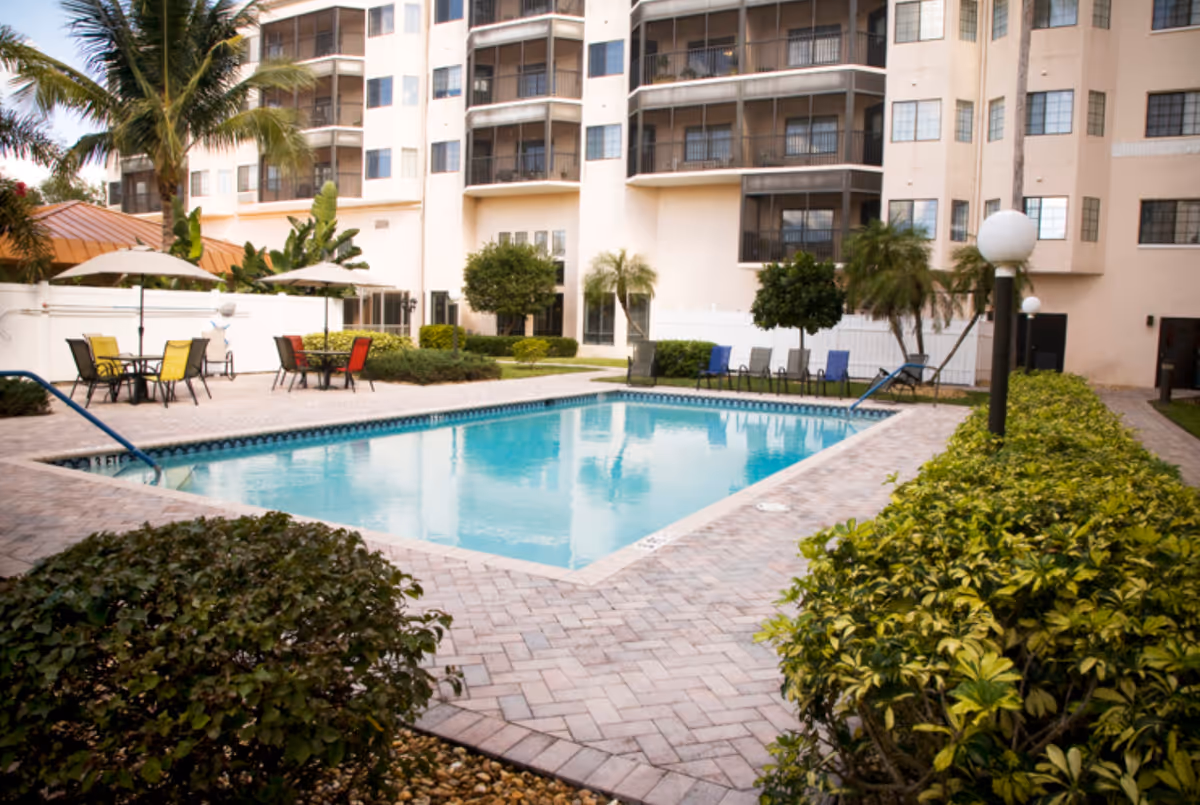 Outdoor swimming pool area at The Palms of Fort Myers with lounge chairs, tables with umbrellas, surrounded by bushes and palm trees, and a multi-story residential building in the background.