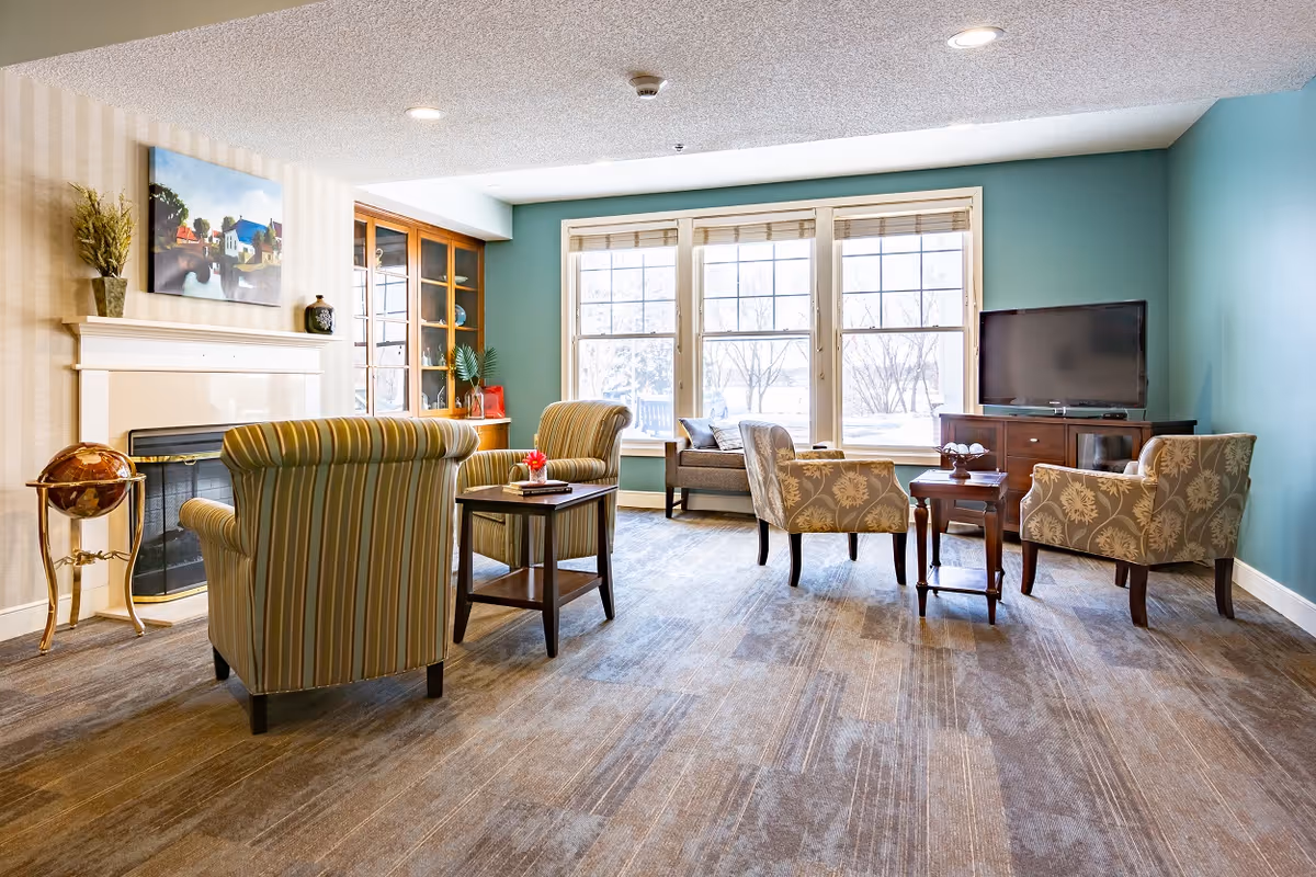 Bright common lounge with upholstered chairs and small tables arranged around a fireplace and TV in front of large windows.
