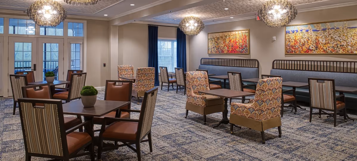 A well-lit dining room with multiple tables and chairs arranged neatly. The chairs have a mix of patterned upholstery and striped backs with brown seats. There are three decorative pendant lights hanging from the ceiling. The room features large windows with blue curtains and two colorful abstract paintings on the wall. The floor is carpeted with a blue and beige pattern.