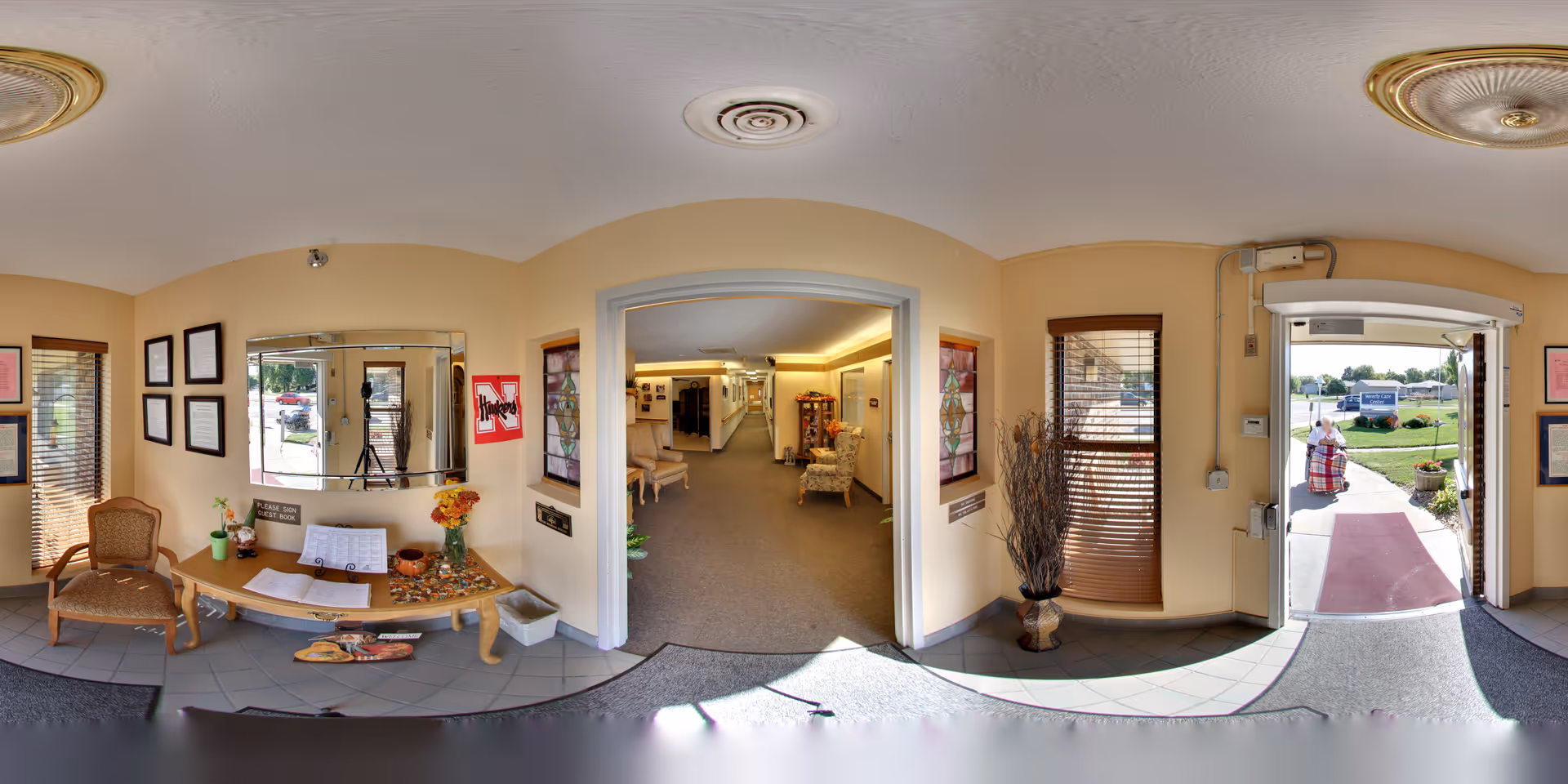 Entrance lobby of a senior living facility with chairs and a table with flowers and decorations, a hallway leading deeper inside, and the exterior visible through an open door.
