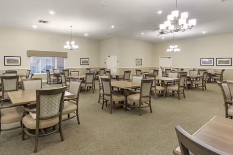 A spacious dining room with multiple wooden tables and cushioned chairs arranged neatly. The room has beige walls, carpeted floors, several framed pictures on the walls, and modern chandeliers hanging from the ceiling providing warm lighting. There are windows with valances allowing natural light into the room.