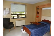 A simple nursing facility bedroom with a single bed covered in a blue blanket, a small overbed table, built-in shelving, an armchair, and a window with a roller shade.