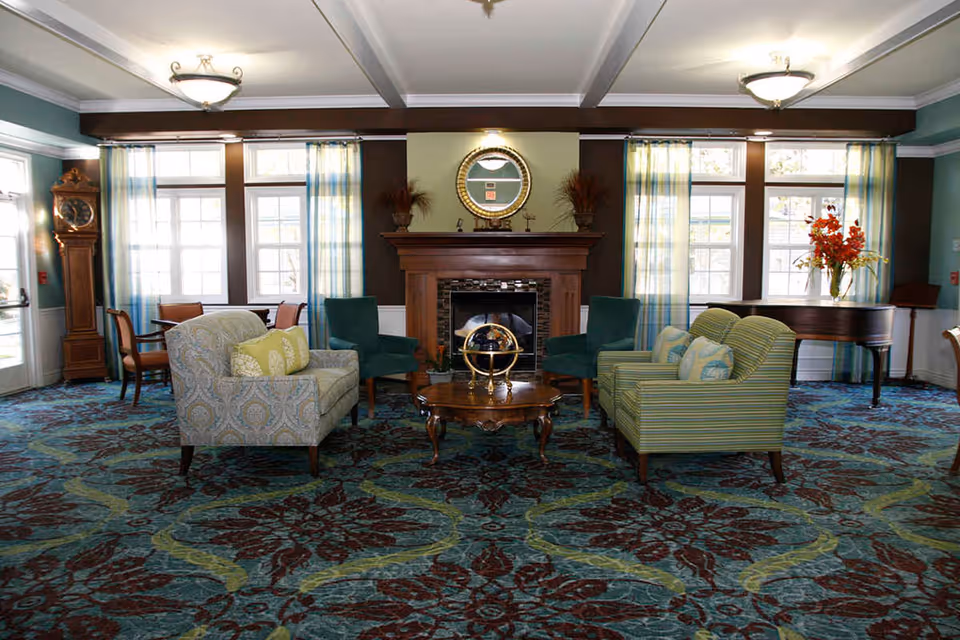 A cozy living room area with patterned carpet, two armchairs, a loveseat, and two green chairs arranged around a wooden coffee table with a decorative globe. The room features a wooden fireplace with a round mirror above it, large windows with sheer curtains, a grandfather clock, and a grand piano with a vase of flowers on top.