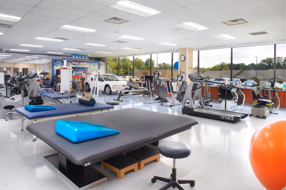 A spacious rehabilitation gym with various exercise equipment including treadmills, stationary bikes, therapy tables with blue wedges, a large orange exercise ball, and a white car used for physical therapy. Large windows provide natural light and a view of trees and buildings outside.