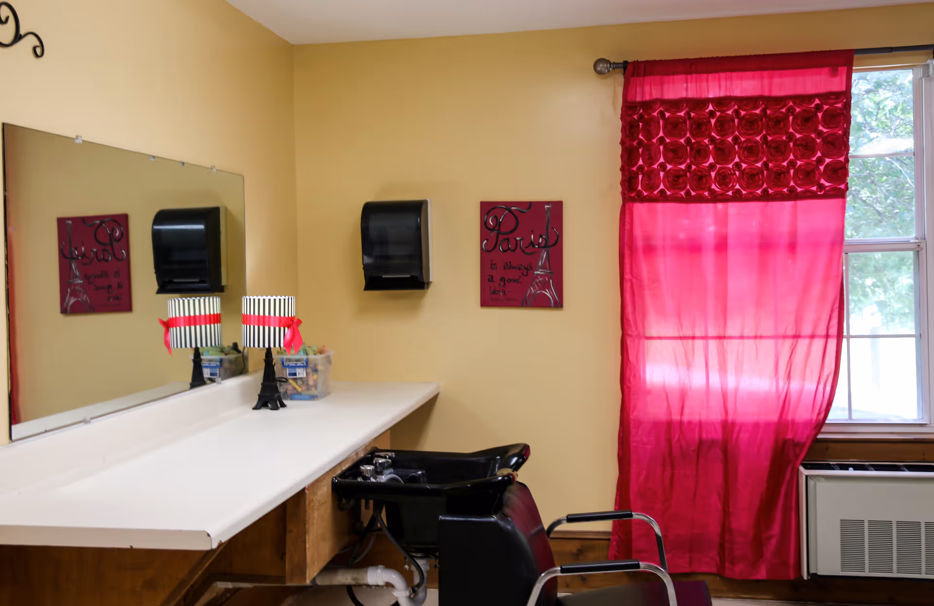 Interior view of a salon area with a black salon chair in front of a white countertop. A large mirror is mounted on the wall above the countertop. There is a black paper towel dispenser on the wall and a decorative sign with the word 'Paris' and an Eiffel Tower illustration. A window with bright pink curtains is on the right side, and an air conditioning unit is below the window.
