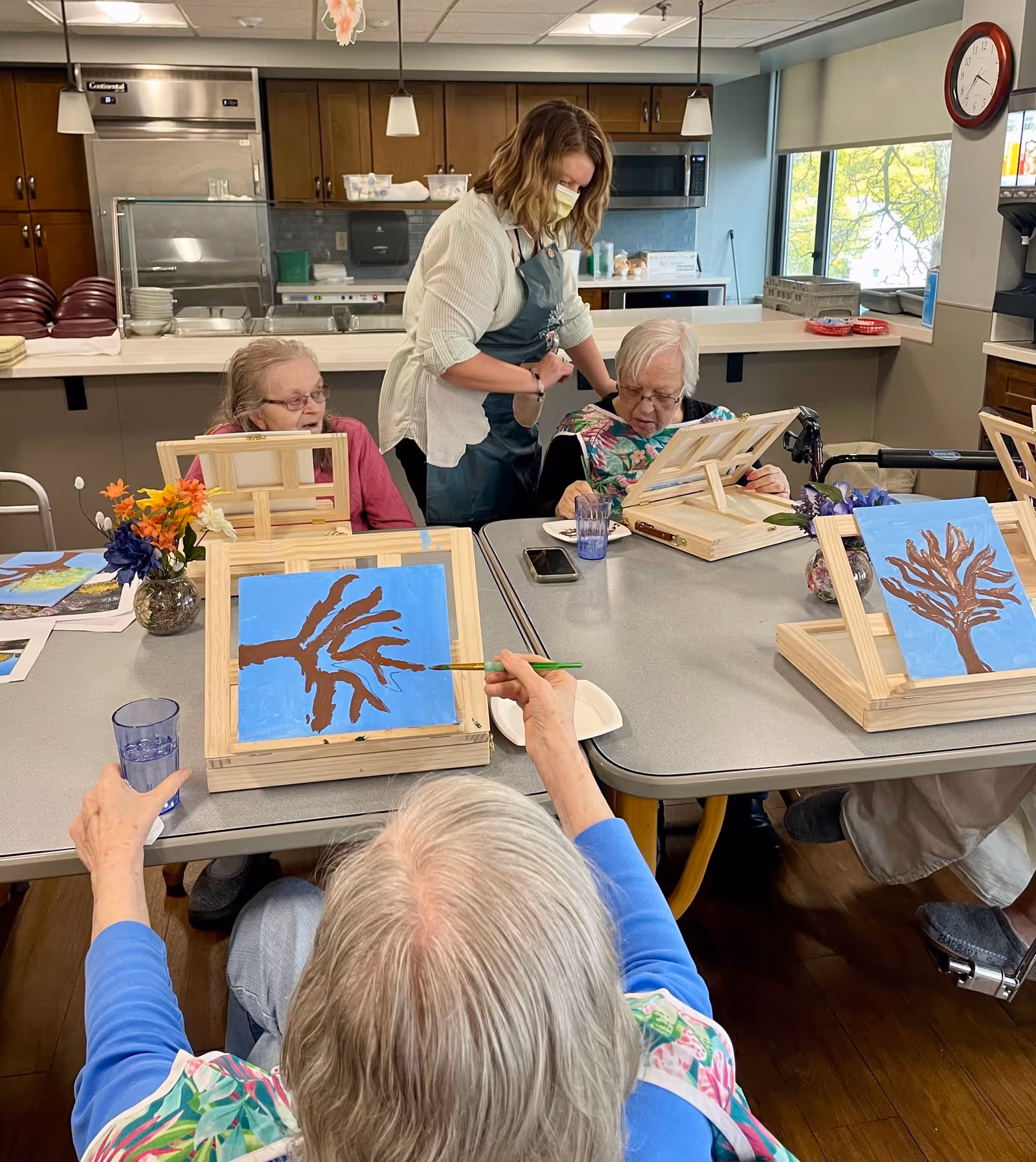 Three elderly women seated around a table in a kitchen or common area, painting pictures of trees on small easels. A caregiver wearing a mask and apron is assisting one of the women. The room has wooden cabinets, a refrigerator, and a clock on the wall.