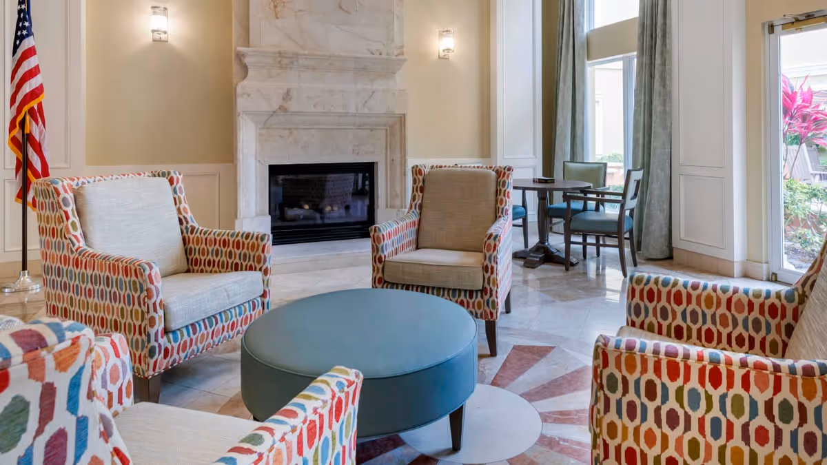 A bright and inviting living room area with four colorful patterned armchairs arranged around a round blue ottoman. The room features a marble fireplace, beige walls with wall sconces, an American flag in the corner, and a small table with chairs near a large window with curtains. Outside, plants and a patio area are visible through a glass door.