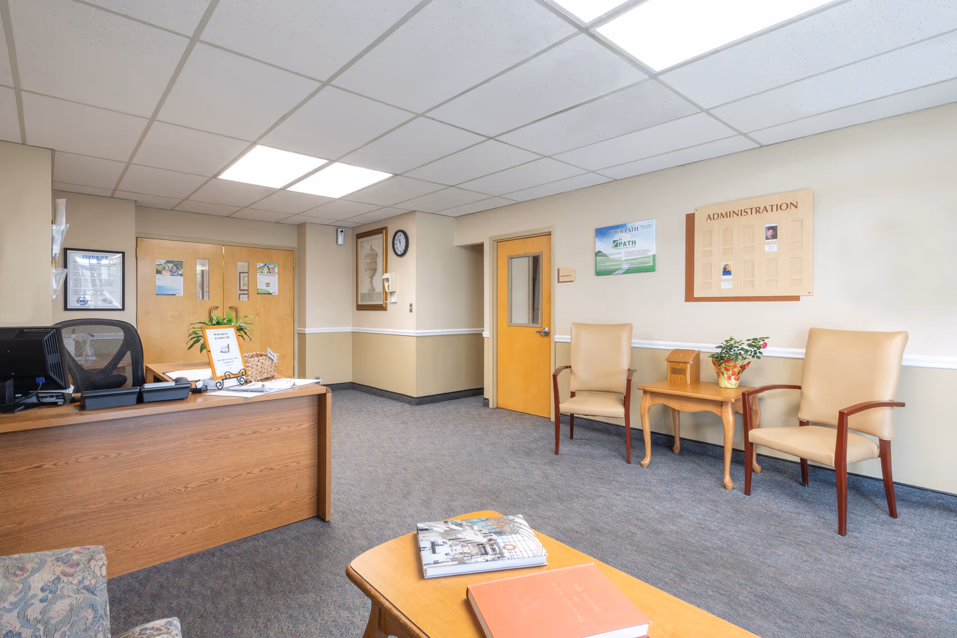 A clean and well-lit reception area with a wooden desk on the left side, two beige chairs with wooden armrests on the right, a small wooden table with a potted plant between the chairs, and a bulletin board labeled 'ADMINISTRATION' on the wall. The room has beige walls with a white chair rail, a gray carpeted floor, and a drop ceiling with fluorescent lights.