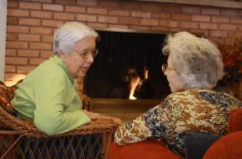 Two elderly women sitting and talking in front of a lit fireplace in a cozy room with a brick wall background.