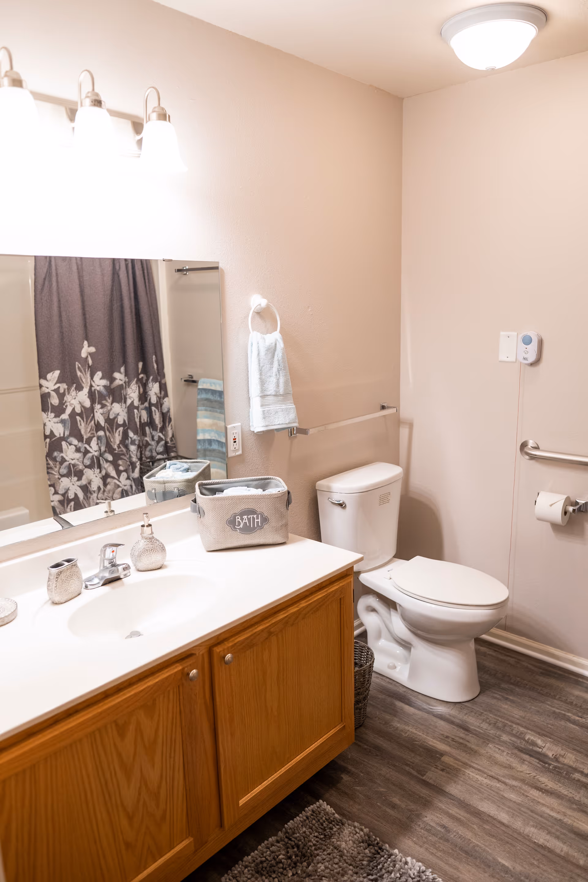 A clean bathroom with a wooden vanity cabinet, white countertop, sink, and faucet. Above the sink is a large mirror with three light fixtures. To the right is a white toilet with a grab bar and toilet paper holder on the wall. A towel ring with a white towel is mounted on the wall next to the mirror. A shower curtain with a floral pattern is visible in the reflection of the mirror. The floor has wood-like vinyl planks and a gray bath mat is on the floor.