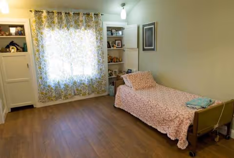 A small bedroom with floral bedspread, window curtains, and built-in shelves.