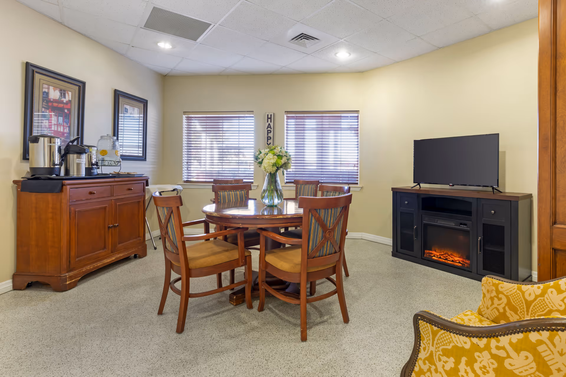 Small communal dining area with a round wooden table and chairs, a sideboard beverage station, and a TV on a cabinet with an electric fireplace.