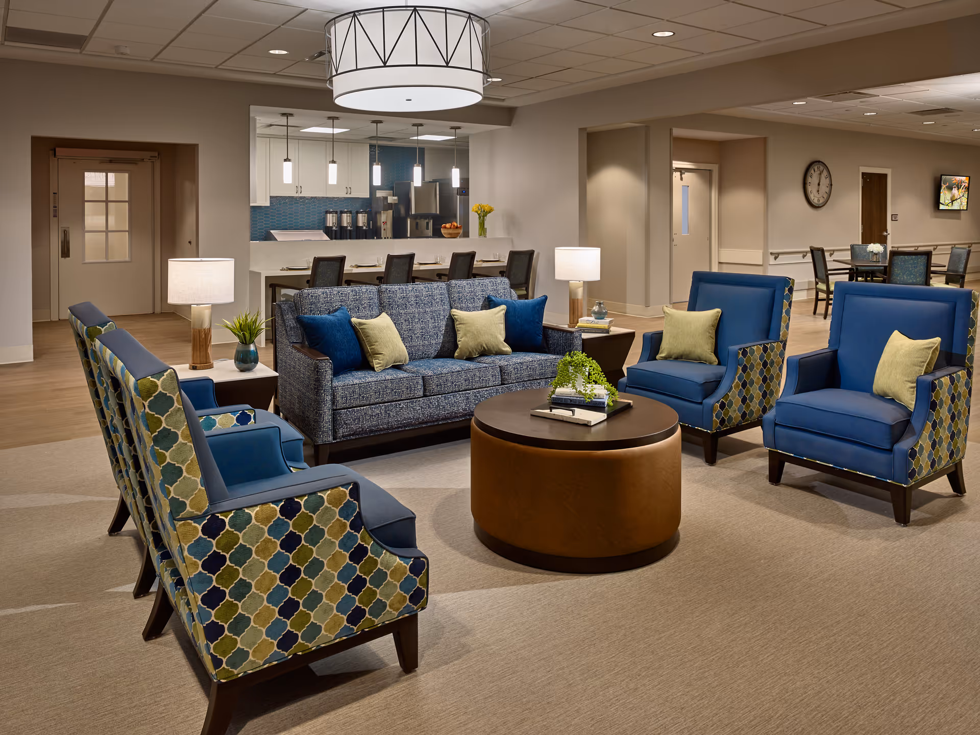 A cozy and modern common area in a senior living facility featuring a blue patterned sofa with green and blue cushions, four blue armchairs with patterned sides, a round brown coffee table with books and a small plant, two side tables with lamps, and a kitchen area with bar stools and pendant lights in the background.