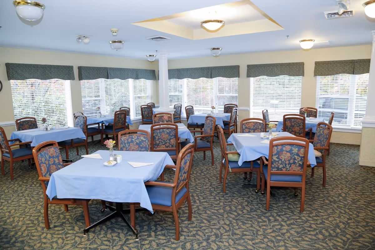 A bright dining room with multiple tables covered in light blue tablecloths, each surrounded by wooden chairs with floral patterned cushions. The room has large windows with blinds and green valances, allowing natural light to fill the space. The carpet has a leafy pattern, and there are ceiling lights and a decorative column in the center of the room.